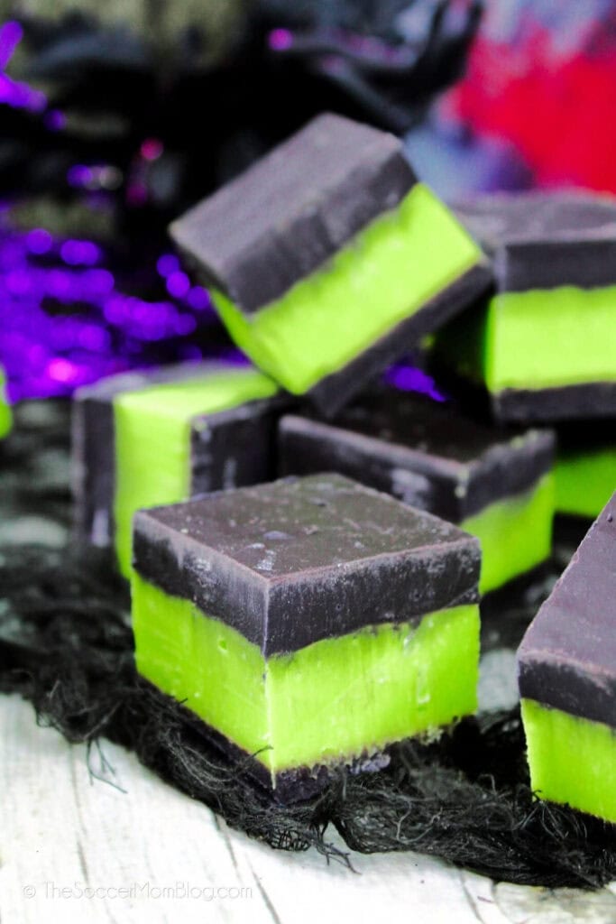 Several square pieces of poison apple fudge have a bright green centre layered between dark chocolate tops and bottoms on a spooky black Halloween cloth.