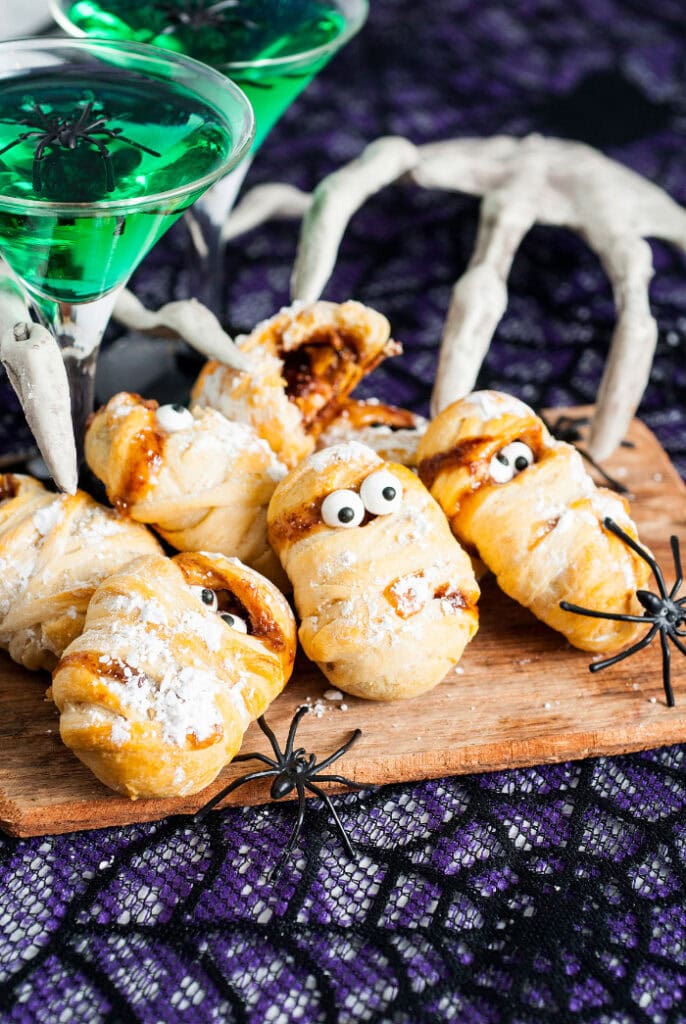 Several golden crescent dough mummy treats with candy eyes sit on a wooden board surrounded by black spiders, skeleton hands, and glasses of green Halloween punch.