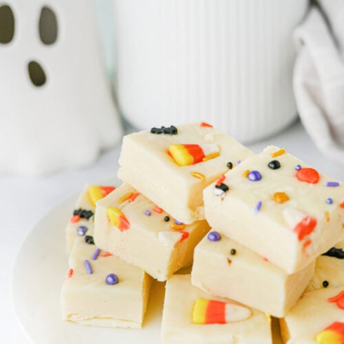 A stack of white chocolate Halloween fudge squares topped with candy corn and colourful sprinkles on a white plate, with a small ghost decoration in the background.