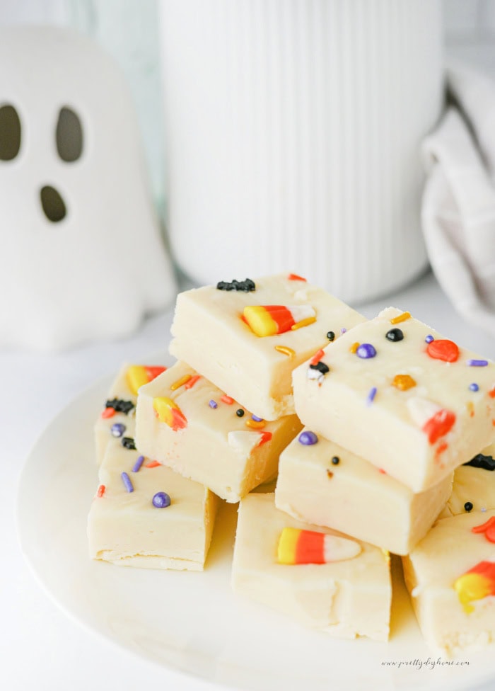 A stack of white chocolate Halloween fudge squares topped with candy corn and colourful sprinkles on a white plate, with a small ghost decoration in the background.
