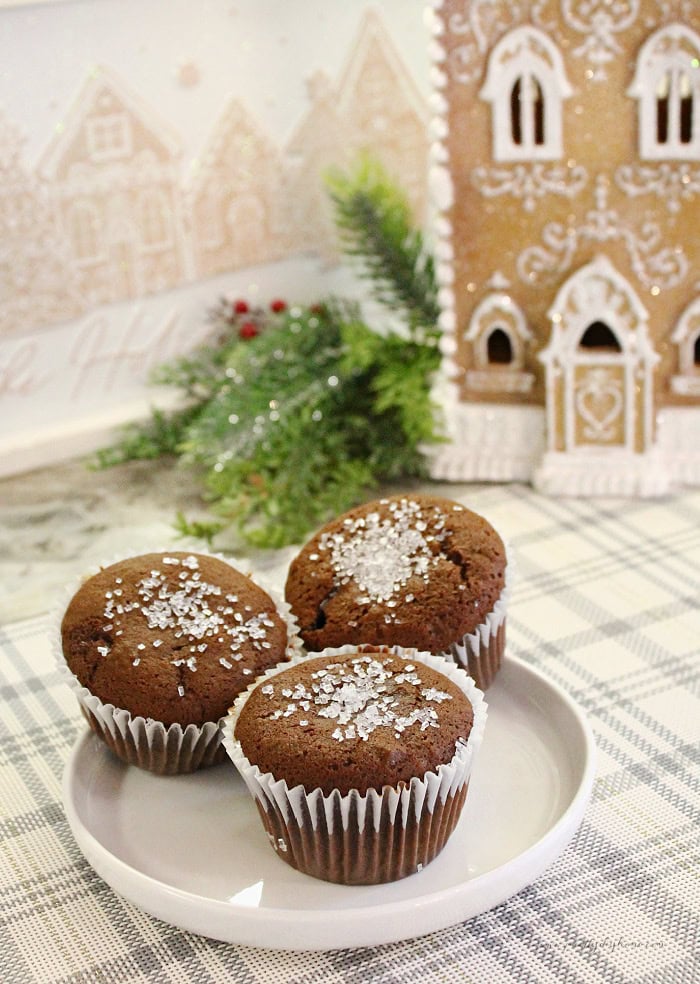 Three gingerbread muffins topped with sparkling sugar beside a gingerbread house and Christmas greenery.