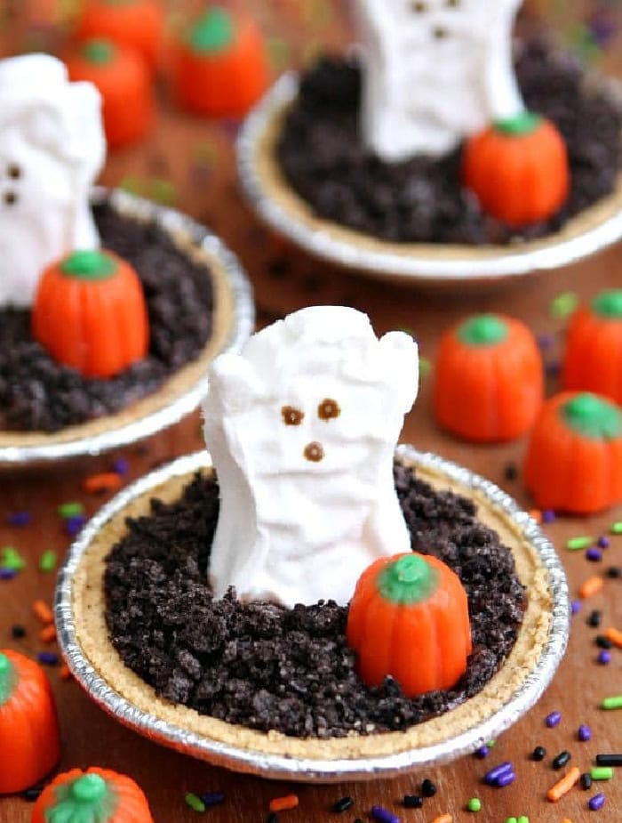 Individual pudding pies in small foil crusts are topped with crushed chocolate cookies, marshmallow ghosts, and orange candy pumpkins.