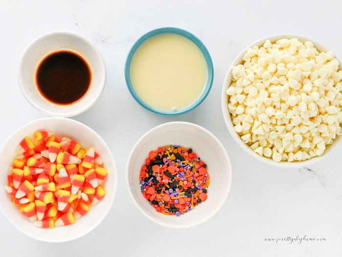 An overhead view of the ingredients for white chocolate Halloween fudge arranged in bowls on a white marble counter. Ingredients include vanilla extract, condensed milk, white chocolate chips, candy corn and Halloween sprinkles.