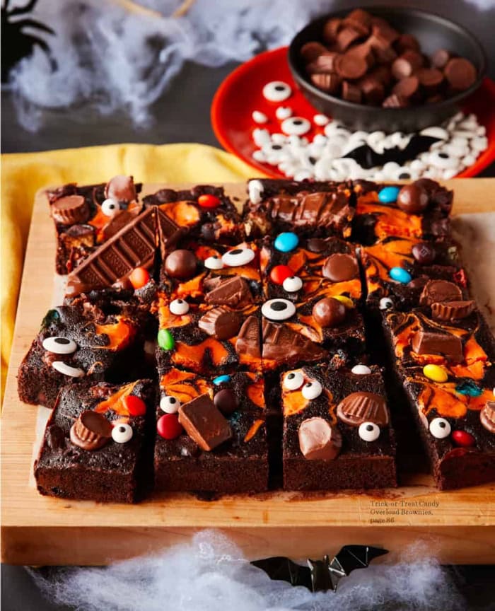 Thick chocolate brownies are topped with orange cream cheese swirls, assorted candy bars, colourful chocolates, and playful candy eyes on a wooden serving board.
