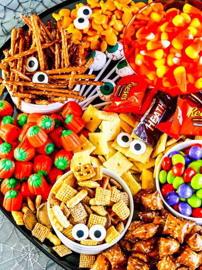 An overhead view shows a large Halloween snack tray filled with pretzels, crackers, cereal mix, candy corn, pumpkin candies, chocolate bars, and colourful sweets decorated with candy eyes.