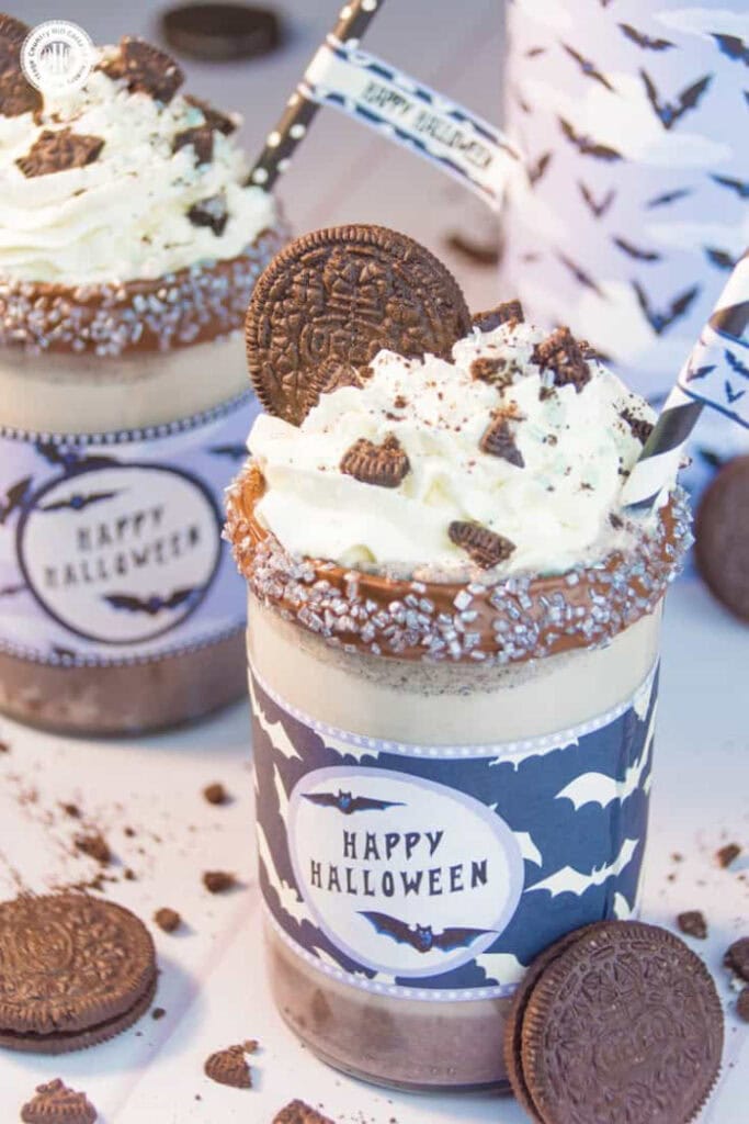 Two creamy Oreo milkshakes are topped with whipped cream, crushed chocolate cookies, and whole Oreos, then decorated with chocolate rims, silver sprinkles, striped straws, and Halloween bat wrappers.