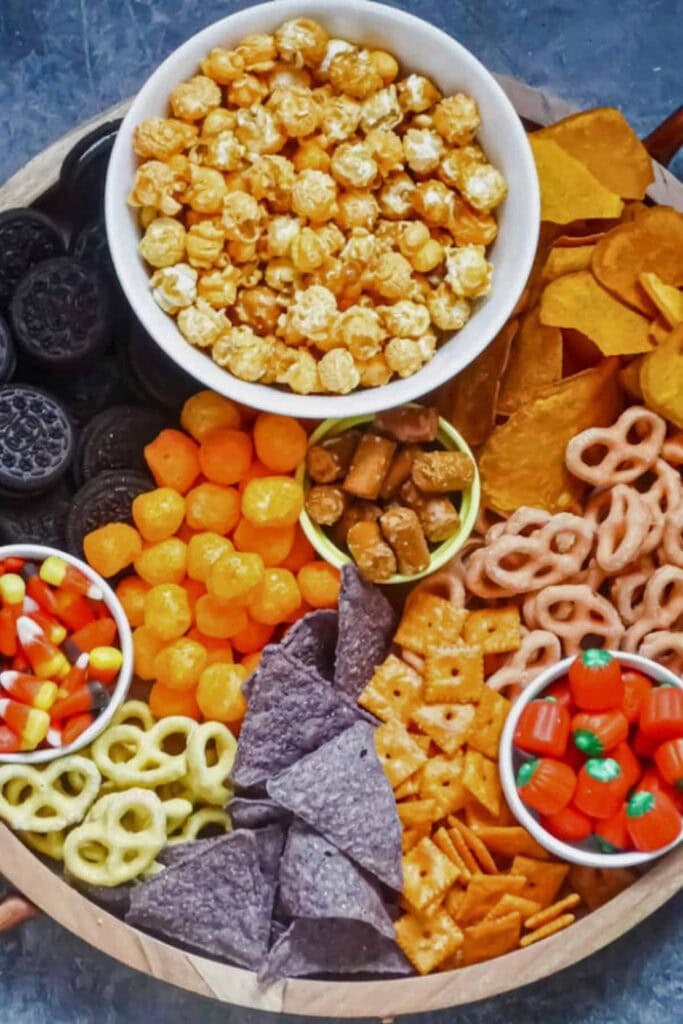 A large round snack board is filled with caramel popcorn, cookies, pretzels, tortilla chips, crackers, cheese puffs, candy corn, and pumpkin shaped candies.