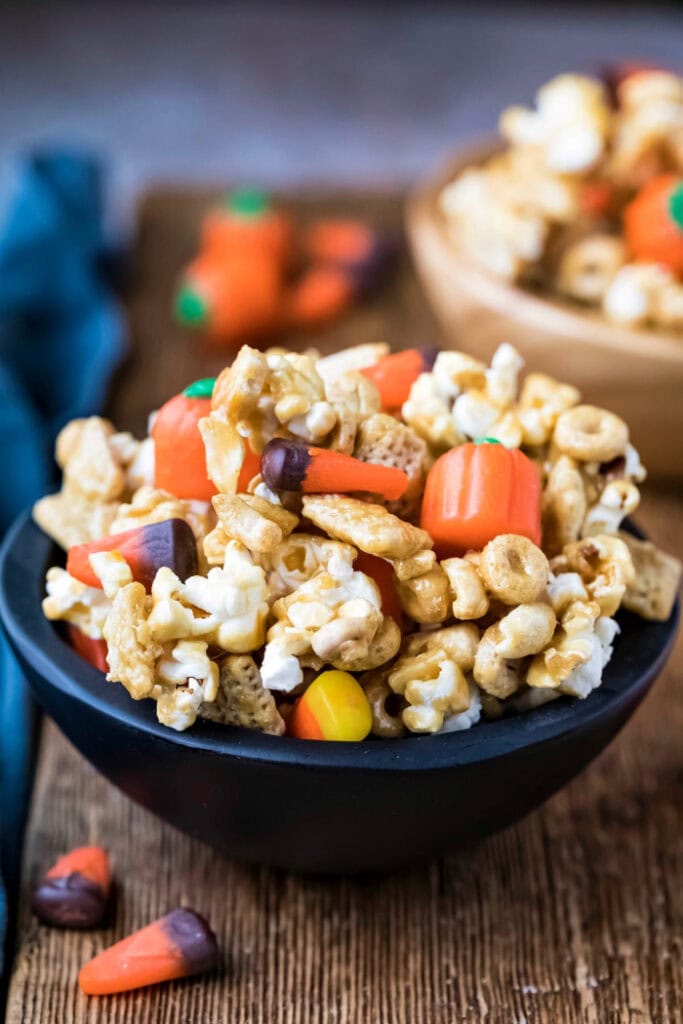 A black bowl is filled with Halloween snack mix made with popcorn, cereal, candy corn, pumpkin candies, and small marshmallows.