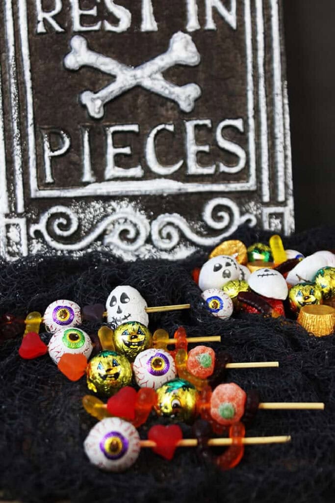 Several wooden skewers are threaded with gummy candies, chocolate eyeballs, foil wrapped pumpkins, skull candies, and other colourful Halloween sweets.