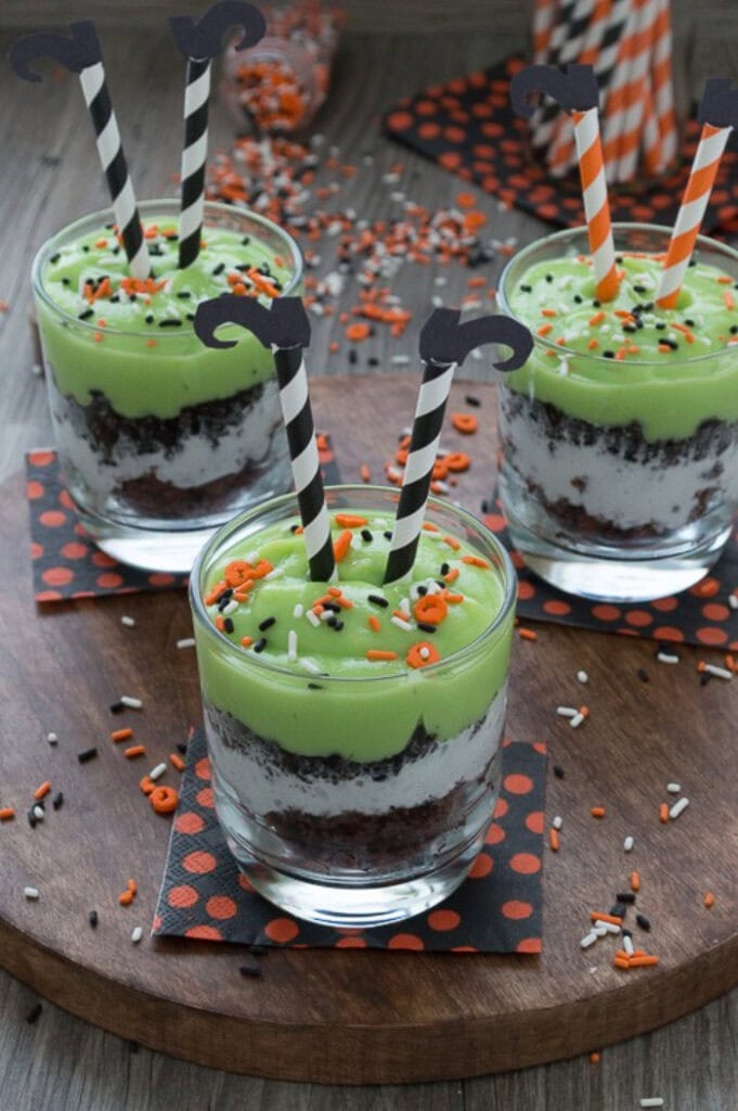 Three melted witch pudding parfaits are layered with chocolate pudding, whipped cream, crushed cookies, and bright green pudding, then topped with Halloween sprinkles and striped witch leg decorations.