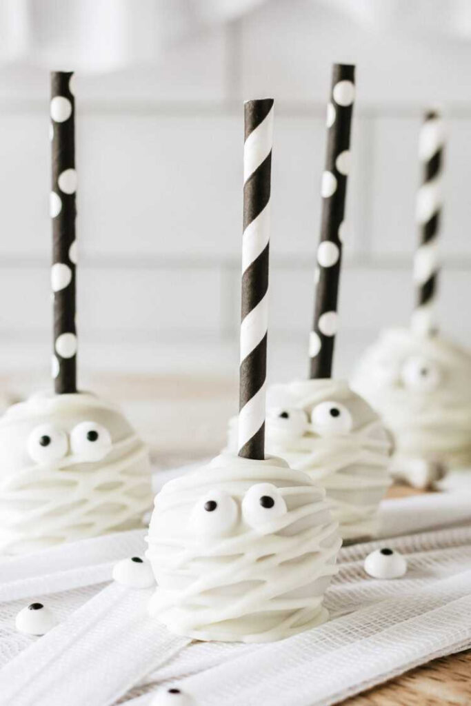 Several round mummy cake pops are coated in white chocolate, drizzled with thin chocolate bandages, and decorated with candy eyes and black and white paper straws.