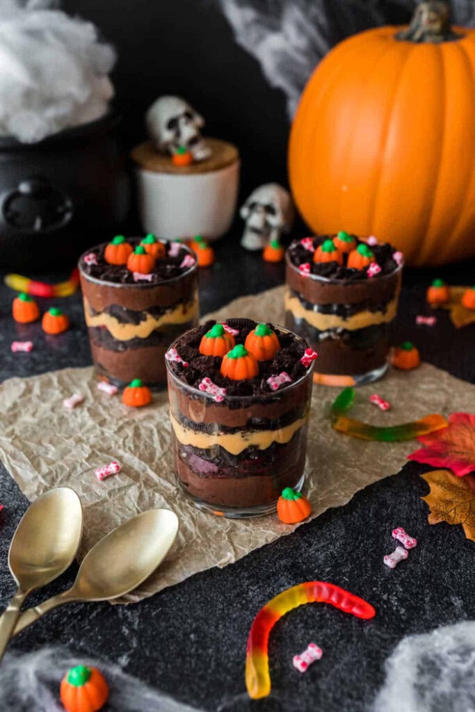 Three clear dessert cups are layered with chocolate pudding, orange filling, and crushed chocolate cookies, then topped with candy pumpkins for a spooky pumpkin patch look.