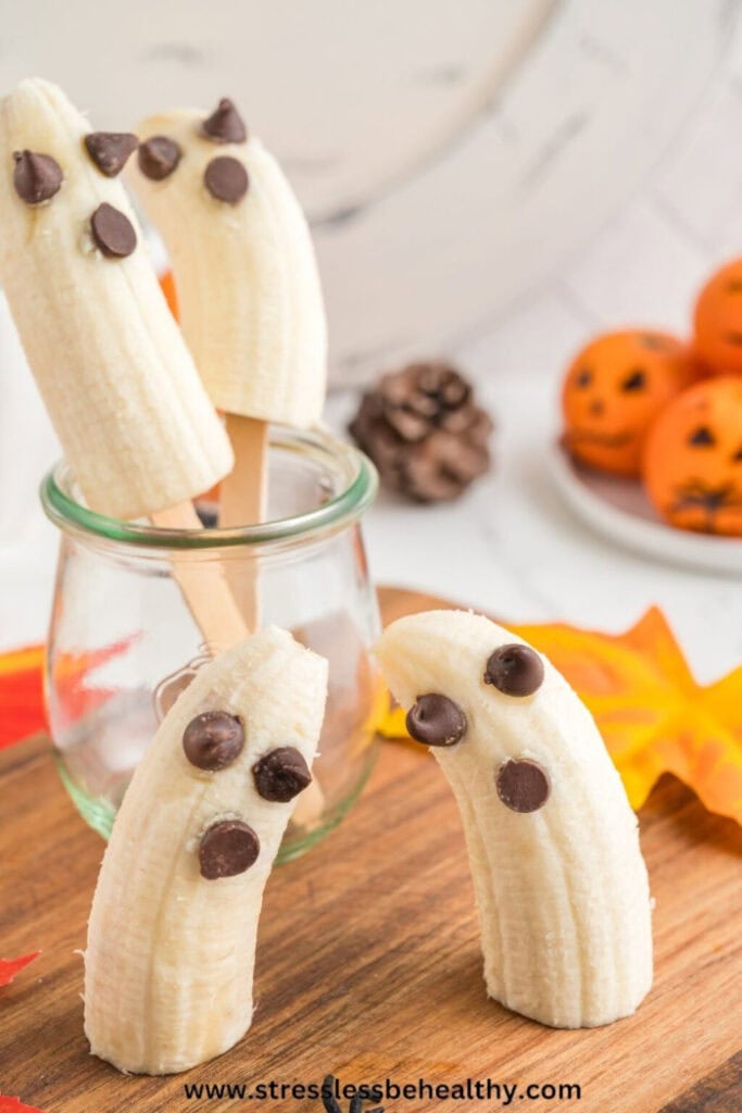 Peeled banana halves are decorated with chocolate chips to create simple ghost faces, with two standing on a wooden board and two displayed on sticks in a glass jar.