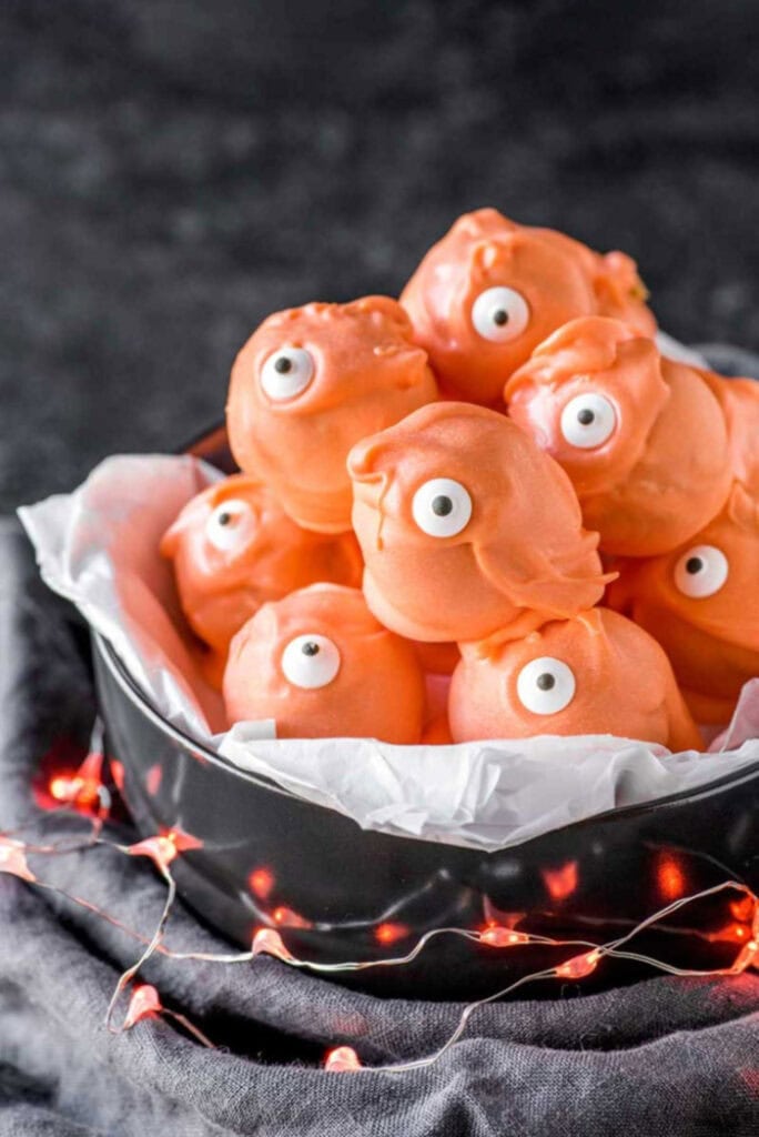 A black bowl lined with white paper holds orange candy coated monster truffles, each decorated with a single candy eye, while orange string lights glow around the bowl.