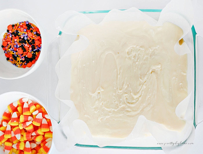 White chocolate Halloween fudge smoothed into a parchment-lined pan with candy corn and sprinkles nearby.