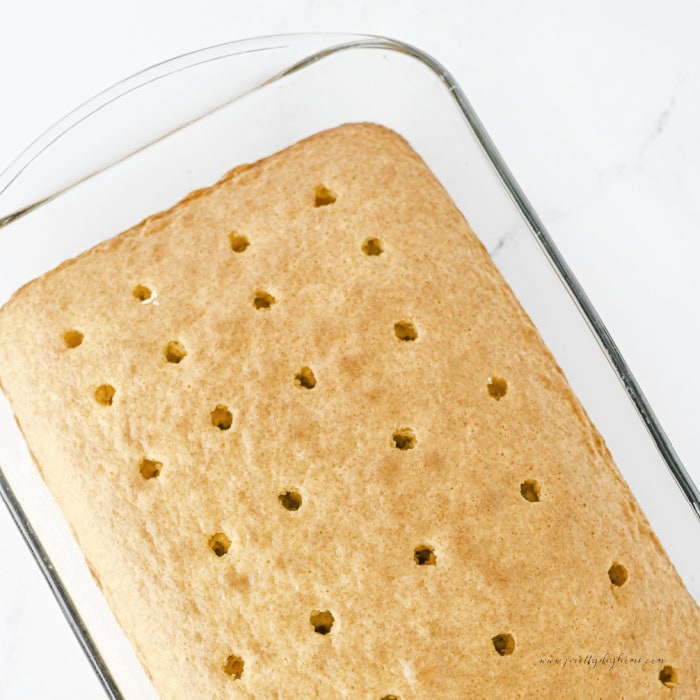 Freshly baked cinnamon roll poke cake in a glass baking dish with evenly spaced holes made across the warm golden cake for the cinnamon filling.