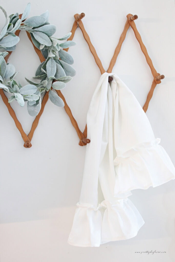 Two finished white damask tea towels with gathered ruffles hanging from a vintage wooden wall rack.