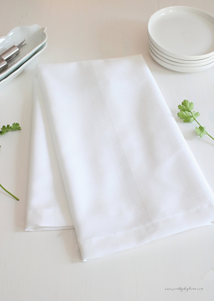 A finished white damask tea towel folded on a kitchen table beside white plates, silverware and fresh cilantro herbs.