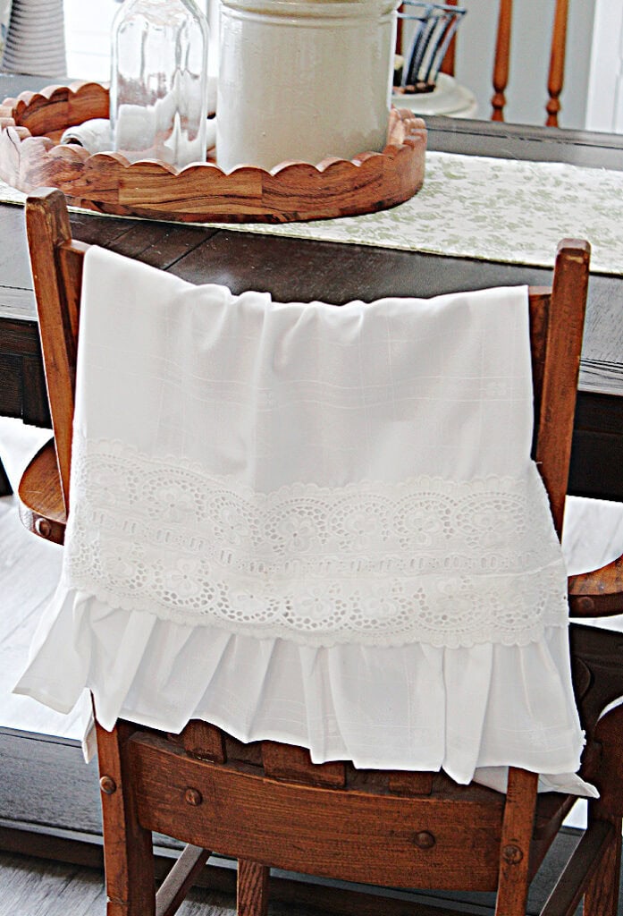 White damask tea towel with a wide lace band and gathered ruffle displayed over an antique wooden chair.