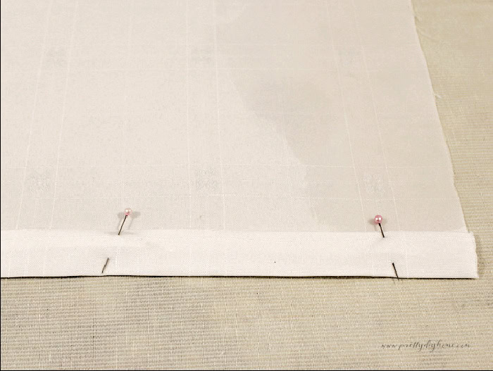 One inch hem folded and pinned along the edge of a white damask tea towel.