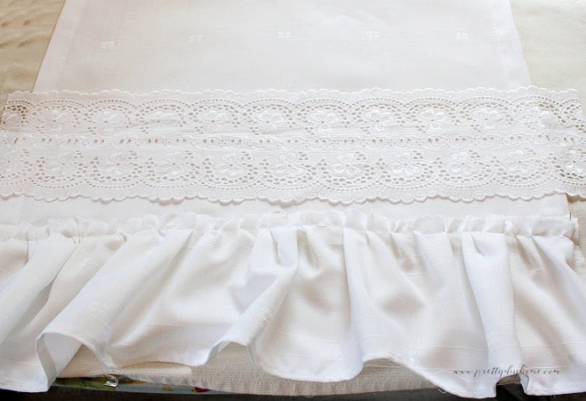 Wide vintage lace ribbon positioned over the sewn edge of a white damask tea towel with an attached ruffle.