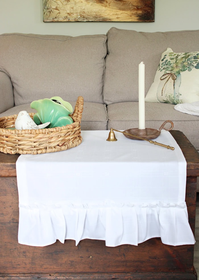 White ruffled damask tea towel used as a small table runner on a rustic wooden coffee table.