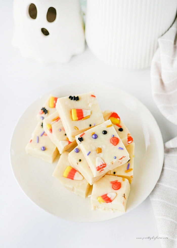 A plate of white chocolate Halloween fudge squares topped with candy corn and colourful sprinkles, with a small ghost decoration in the background.