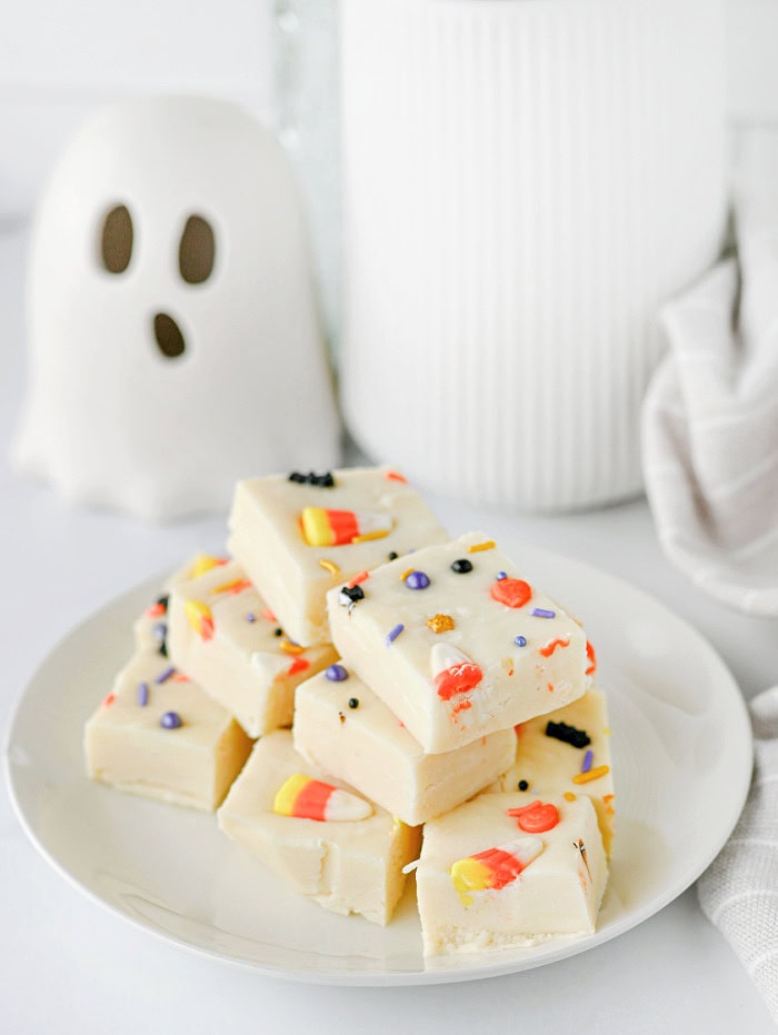 A plate of white chocolate Halloween fudge squares topped with candy corn and sprinkles, with a small ghost decoration in the background.