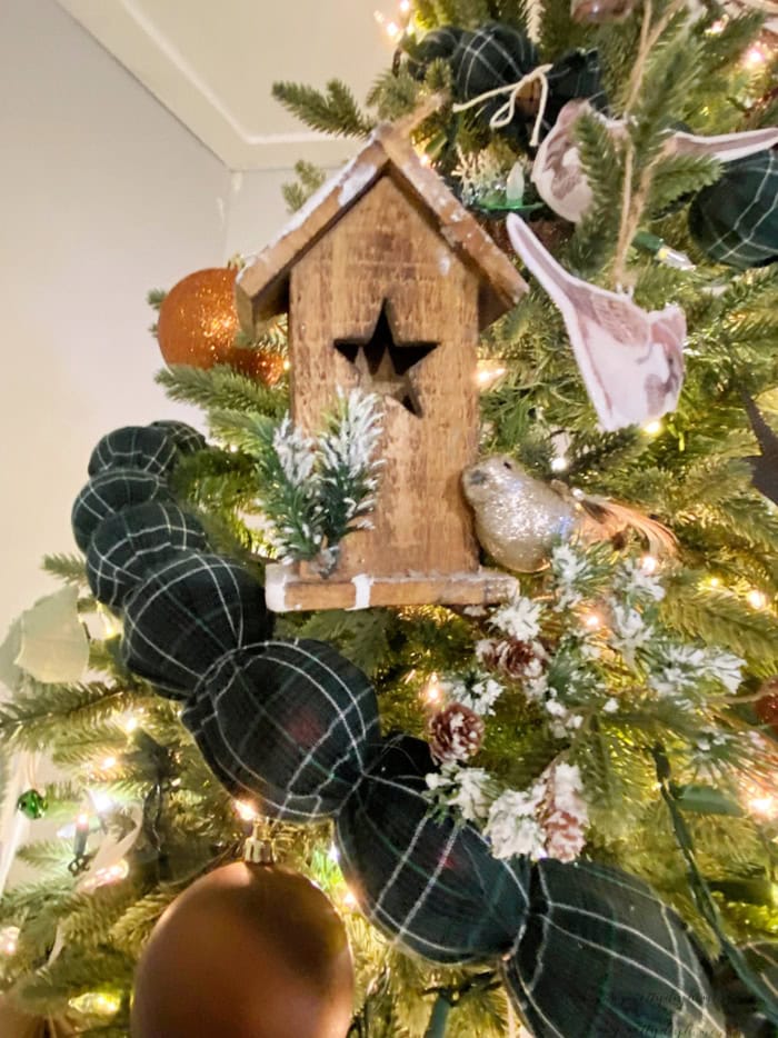 Green tartan fabric garland draped across a Christmas tree with a rustic wooden birdhouse, greenery, and ornaments.
