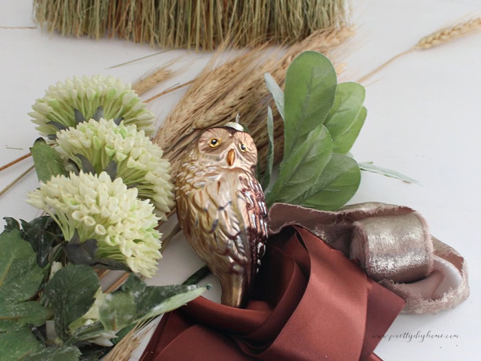 Supplies for a decorated cinnamon broom including faux greenery, wheat, cream flowers, ribbon, and a small owl ornament.