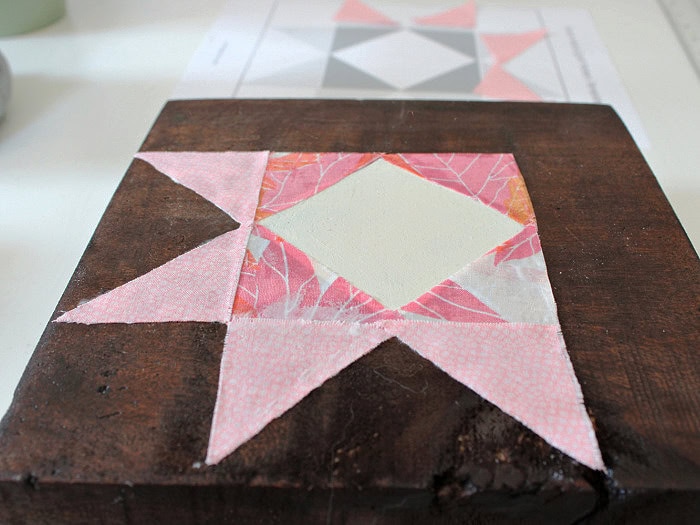 Pink fabric triangles being added around the leaf-print center pieces on a stained wooden barn quilt block.