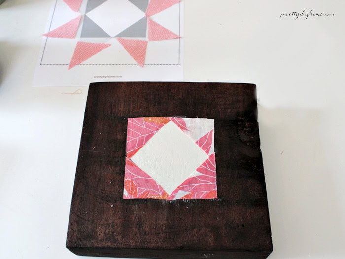 Four leaf-print fabric pieces attached around the cream center diamond on a stained wooden barn quilt block.