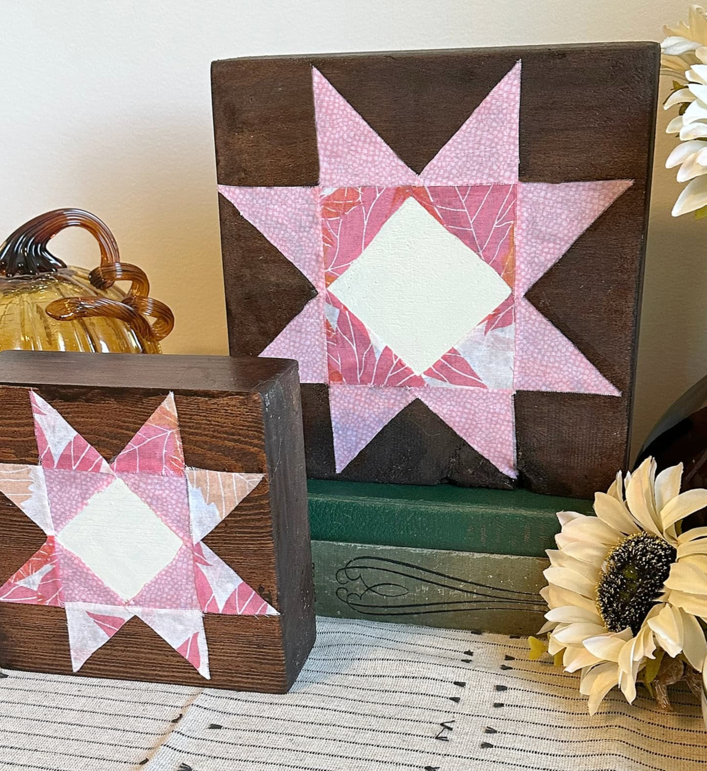 Close-up of two pink fabric barn quilt shelf sitters displayed on vintage books with cream flowers.