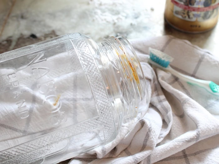 A vintage glass jar with rust around the rim lies on a towel beside a toothbrush and cleaner before the rust is scrubbed away.