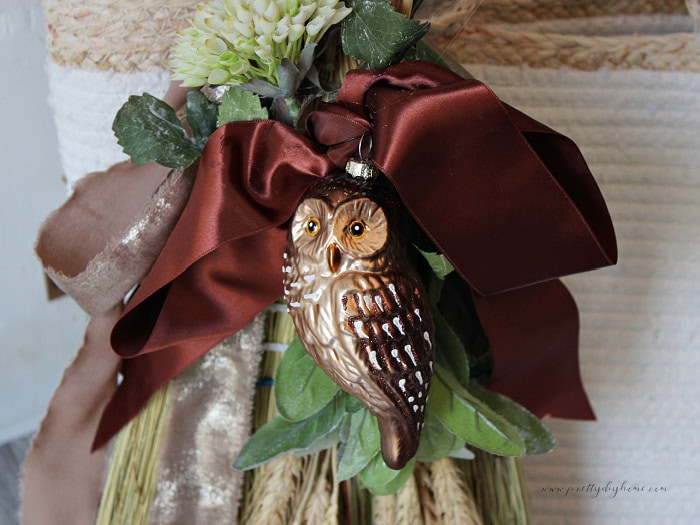 Close-up of a decorated cinnamon broom with an owl ornament, chocolate satin ribbon, greenery, wheat, and cream flowers.