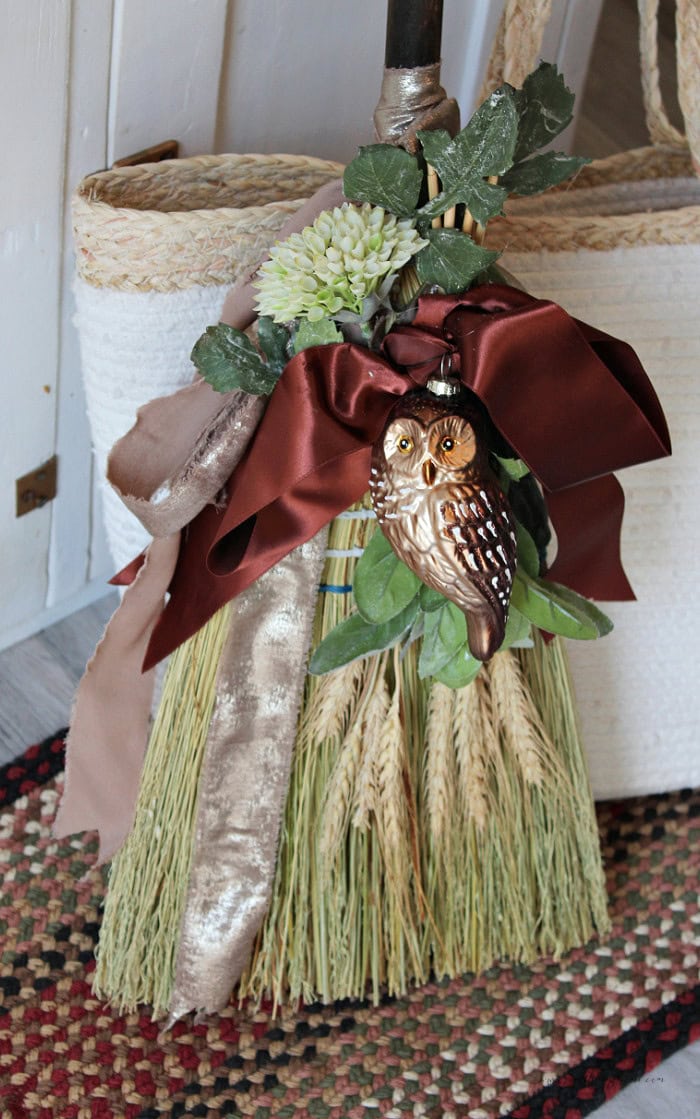 Decorated cinnamon broom with an owl ornament, burgundy ribbon, wheat, and faux greenery for fall.