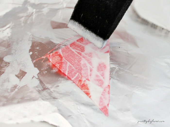 Mod Podge being dabbed onto the back of a pink fabric triangle with a foam brush.