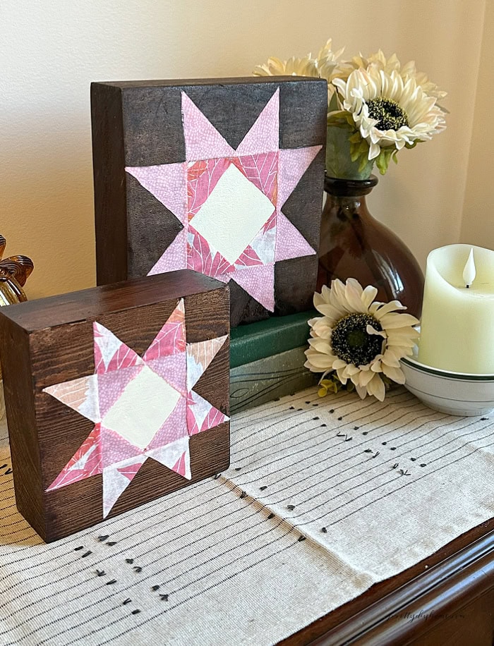 Two pink fabric barn quilt shelf sitters displayed with cream flowers, vintage books, and a candle for fall.