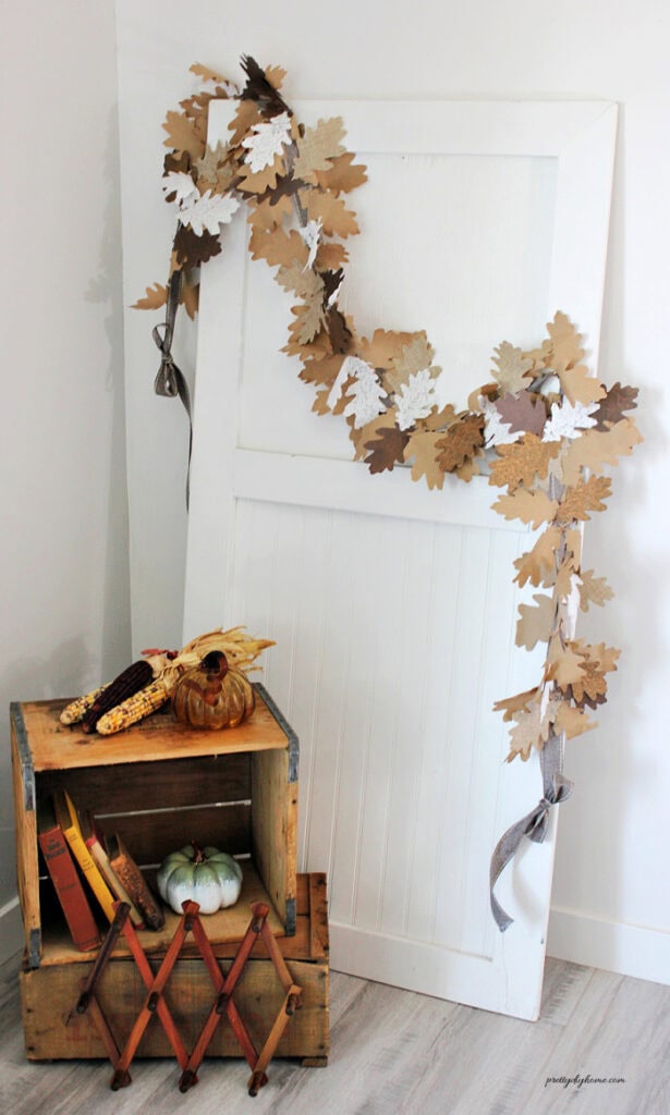This finished fall garland DIY is draped over a white vintage door and styled with wooden crates, pumpkins, old books, and dried corn.