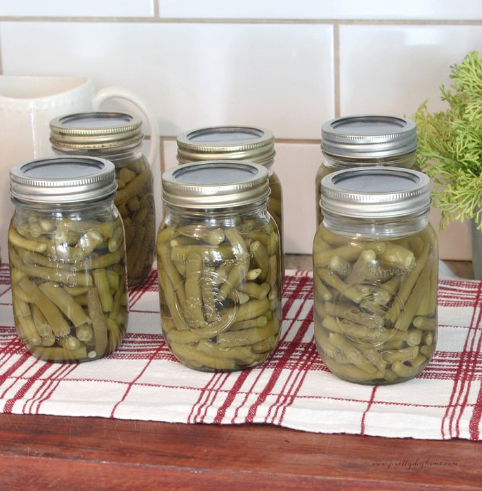 Finished jars of home canned green beans are lined up on a wooden board.