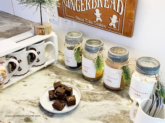 Hot chocolate station with labeled mason jars, gingerbread mugs, chocolate treats, and Christmas greenery on a kitchen counter.