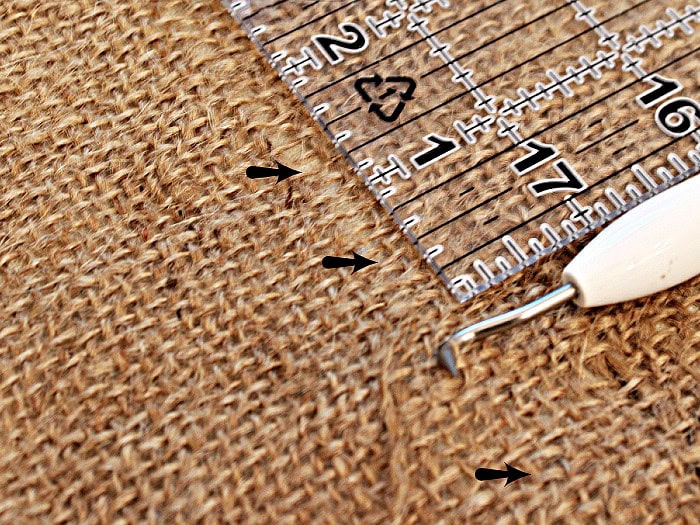 A crochet hook pulling a thread from burlap fabric to create a straight cutting line.