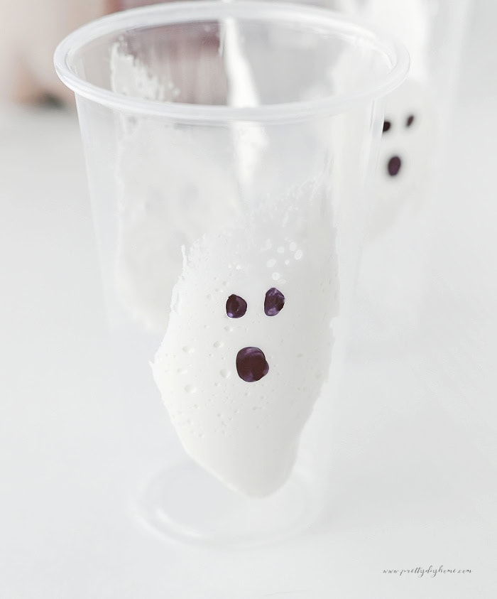 Marshmallow fluff ghost spread inside the side of a clear cup with a Sharpie face drawn on the outside.