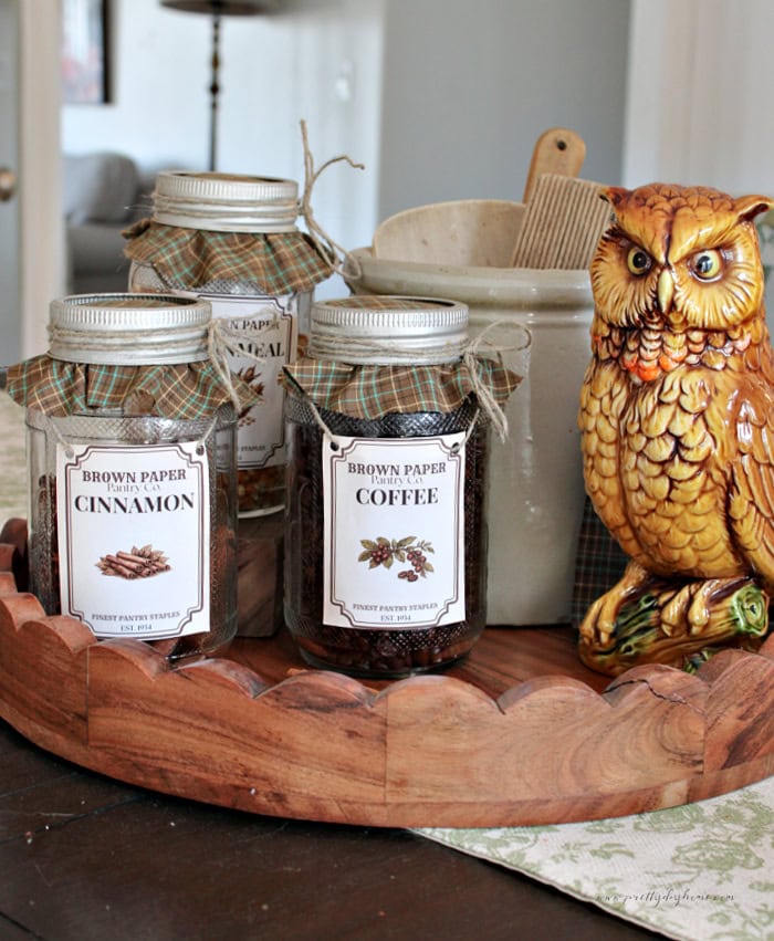 Vintage pantry jars with cinnamon, coffee, and cornmeal labels are arranged on a wooden tray with a ceramic owl for fall decor.