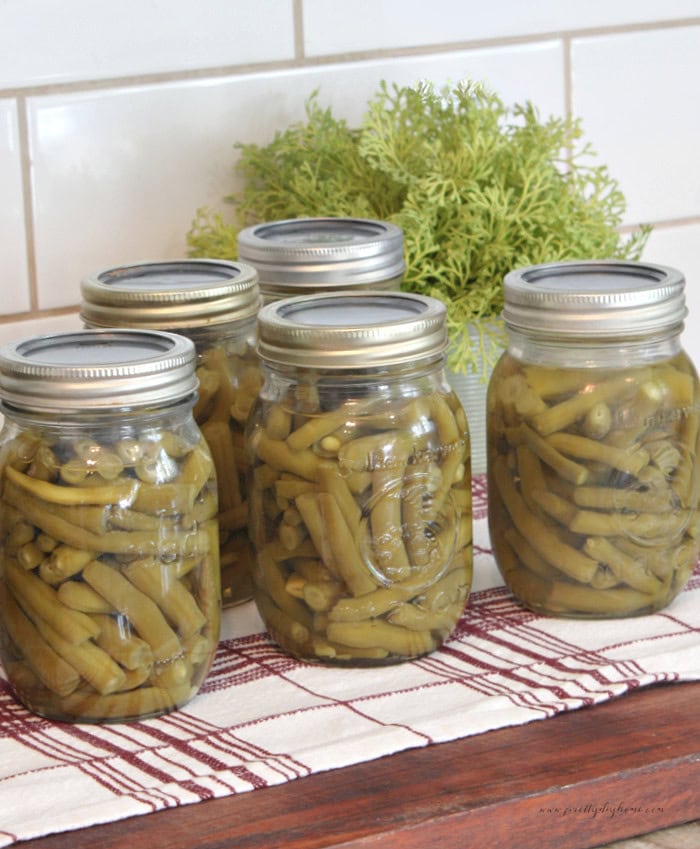 Five jars of pressure canned green beans are lined up on a wooden board and tea towel after processing.