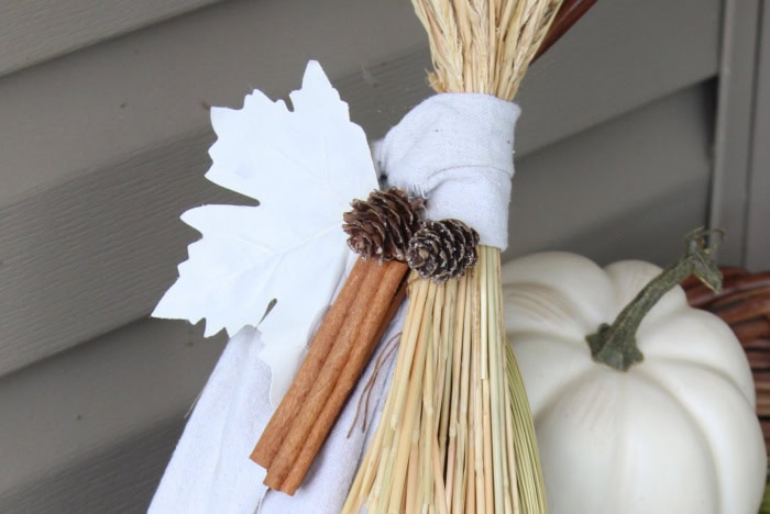 Close-up of primitive cinnamon broom decor with warm white fabric, cinnamon sticks, pinecones, and a white fall leaf.