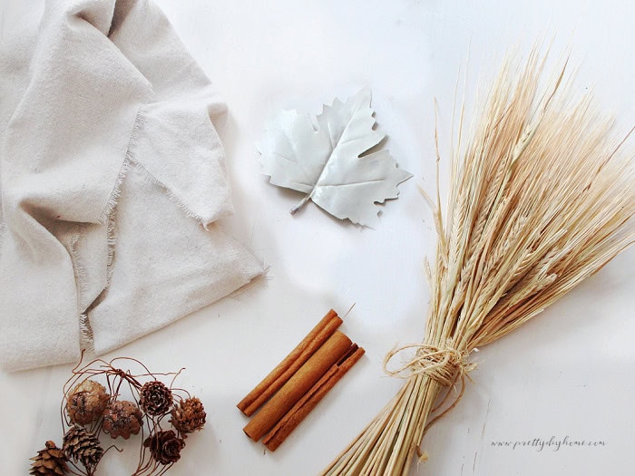 Supplies for primitive cinnamon broom decor including dried wheat, cinnamon sticks, warm white fabric, pinecones, and a white fall leaf.