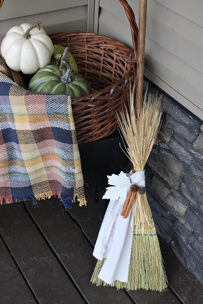 Primitive cinnamon broom fall porch decor styled with a basket, pumpkins, plaid throw, dried wheat, cinnamon sticks, and warm white fabric.