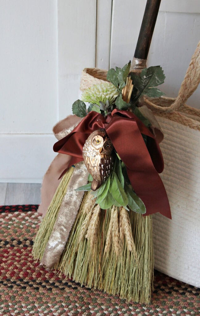 Rustic cinnamon broom with an owl ornament, burgundy ribbon, wheat, greenery, and cream floral accents.