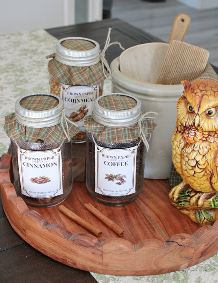 Three vintage pantry jars with cinnamon, coffee, and cornmeal labels are styled on a wooden tray with plaid fabric lids, cinnamon sticks, a crock, and a ceramic owl.